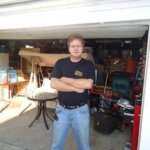 A man with short hair stands with arms crossed in front of an open garage filled with various items, showcasing the need for junk removal in Clifton NJ. He is wearing blue jeans and a dark t-shirt.