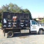 A white and black dump truck with business advertising on the side is parked in a lot near trees and a fence. The door displays contact info for junk removal in Morris County NJ, and the truck bed holds various tools.