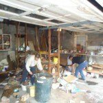 Two people are cleaning and organizing a cluttered garage filled with various items, boxes, and tools—shelves and wooden beams are visible along the walls—showcasing the need for junk removal in Passaic County NJ.