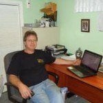 A person sits at a wooden desk in a home office in Passaic County NJ, using a laptop. Office supplies and papers are on shelves and the desk—perfect for someone considering junk removal in Passaic County NJ to clear clutter. Pale green walls display framed pictures.