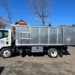 A silver dump truck with a "Milex Carting" logo, specializing in junk removal in Essex County NJ, is parked on a dirt lot. Houses, trees, and other vehicles are in the background, while the truck's bin displays capacity and weight information.