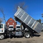 A white dump truck for junk removal in Passaic County NJ with a raised metal bed is parked on a lot. Several white vans and a red house are visible in the background under a clear blue sky.