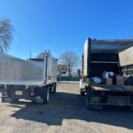 Two trucks for junk removal in Morris County NJ are parked side by side on a sunny day—one with an empty metallic bed, the other loaded with boxes and bags. Leafless trees and houses stand in the background.